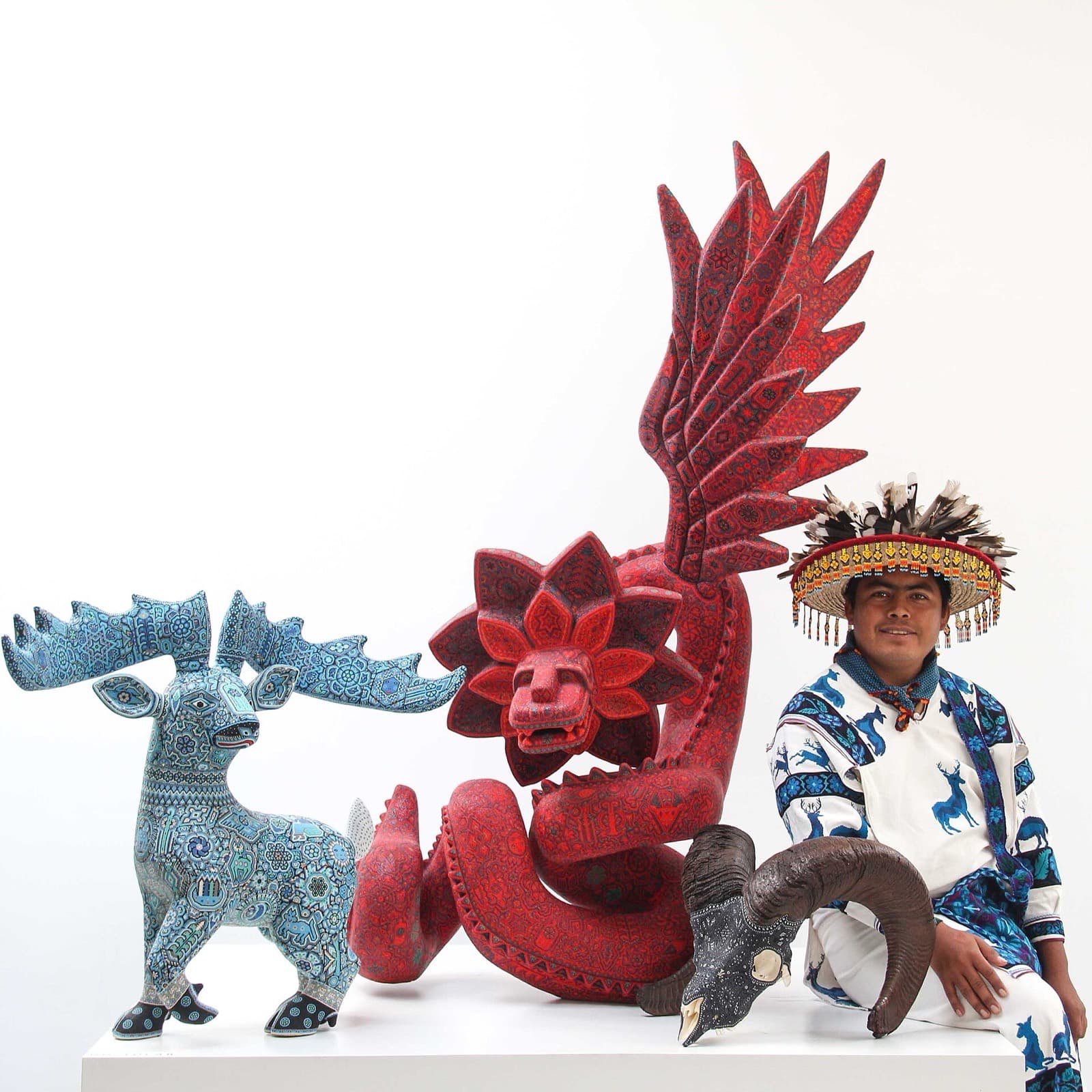 Artista wixárika con sombrero ceremonial de plumas junto a tres esculturas cubiertas de chaquira de cristal