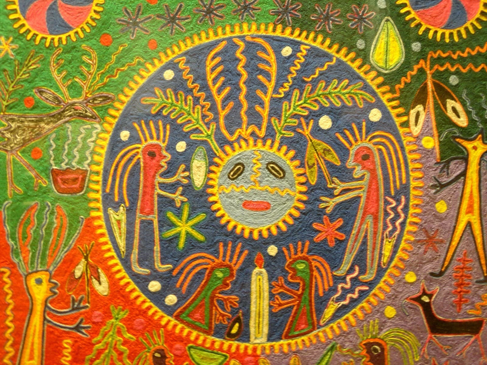Cuadro de estambre huichol con un disco solar de rostro humano rodeado de figuras ceremoniales, venados y velas