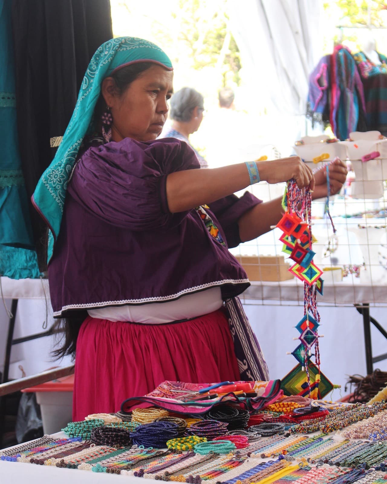 Mujer wixárika con indumentaria tradicional y rebozo turquesa mostrando ojos de dios y collares de chaquira en un tianguis artesanal